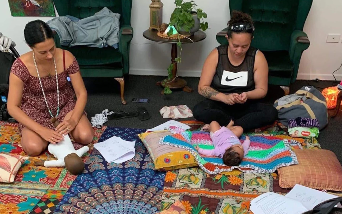 Two women demonstrate how to massage babies. The babies lie on coloured blankets on the floor.