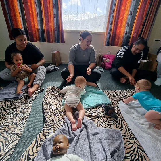 Three smiling women sit on the floor with their babies on blankets in front of them.